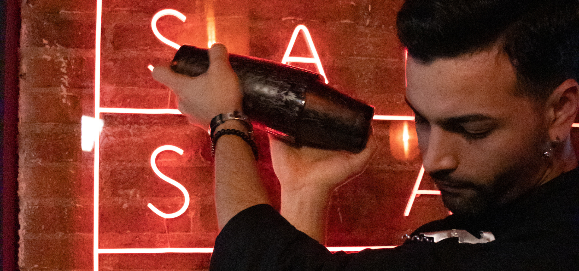 Bartender preparando un cóctel con coctelera metálica frente a un neón rojo con letras en Maemuki